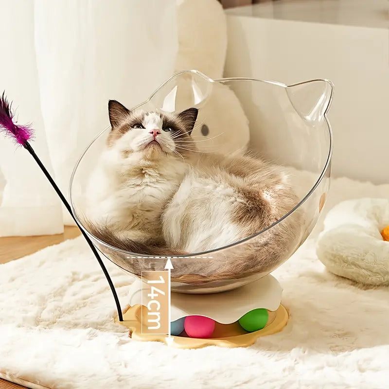 A cat sitting inside an acrylic capsule-shaped cat bowl, which is placed on a white surface with a toy wand and a track ball next to it with measurements 