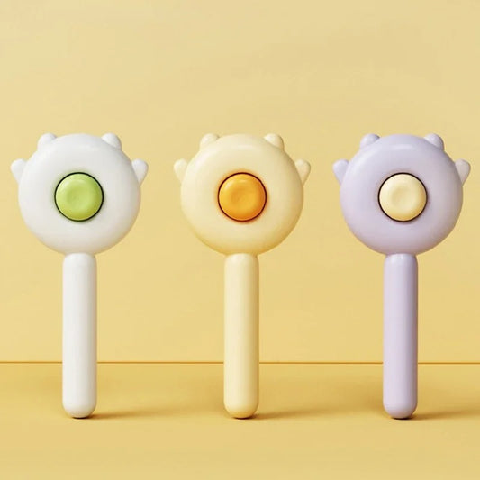 Three cat hair brushes with different colors (green, yellow, and purple) each featuring a stylized cat face at the brush head, displayed against a light yellow background.