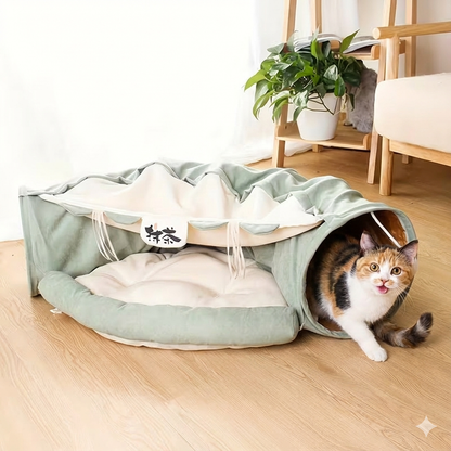 Cat peeking out from a green pet bed on a wooden floor with a plant and chair in the background.