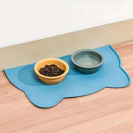A blue silicone feeding mat with raised edges, placed on a wooden floor under two bowls, one containing cat food and the other filled with water.