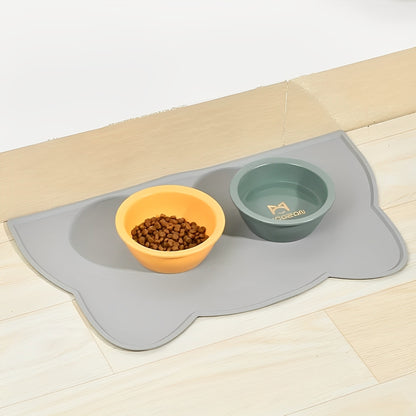 A gray silicone feeding mat with raised edges, placed on a wooden floor under two bowls, one containing cat food and the other filled with water.