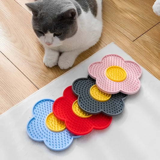 A cat sitting next to a slow feeder mat licking pad with suction cups. The mat has a floral pattern with pink, blue, grey and redflowers.